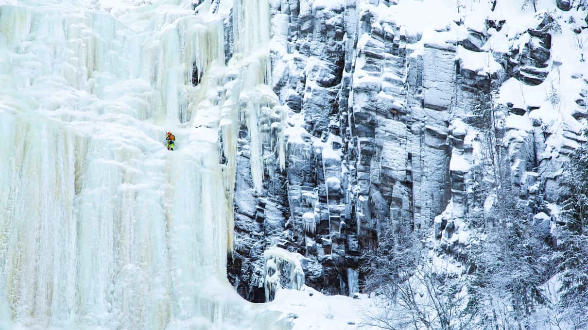 A tall column of white ice beside a dark frost-covered cliff, with a tiny climber halfway up