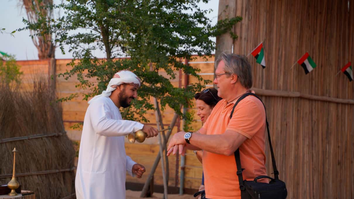 Un uomo in kandura bianca offre il caffè arabo da una dallah a due ospiti, sul recinto di canne le bandierine degli Emirati