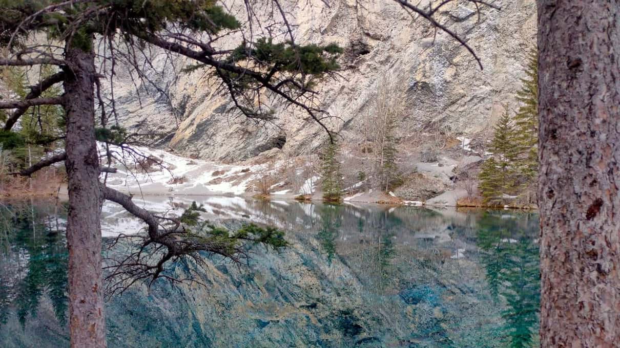 A small lake with patches of snow on its shore, reflecting a pale limestone cliff behind tall pines