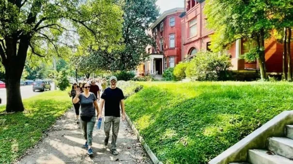 A small group walking along a sunny path through a green park toward red-brick houses in Old Louisville