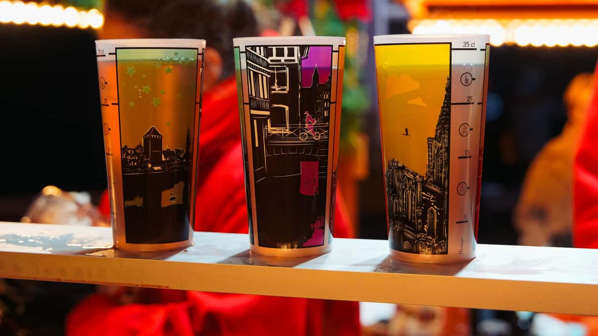 Three steaming mugs of mulled wine printed with Strasbourg designs lined up on a market counter