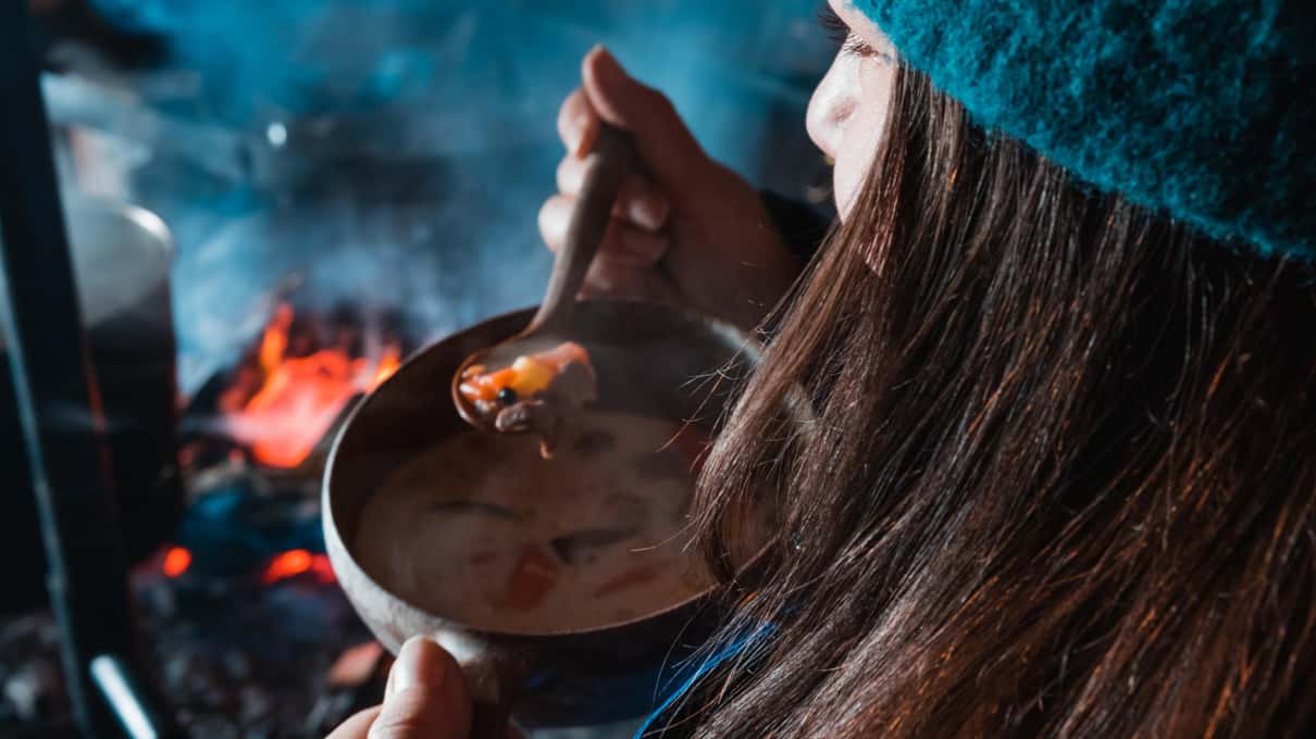 A woman in a teal knit hat eating hot soup beside a campfire on a Safartica ice fishing trip