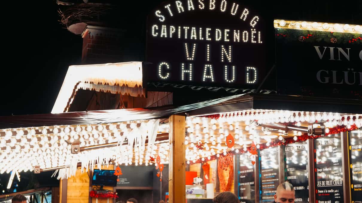 A brightly lit market stall at night under a sign reading Capitale de Noël, with Vin chaud lettered above the counter