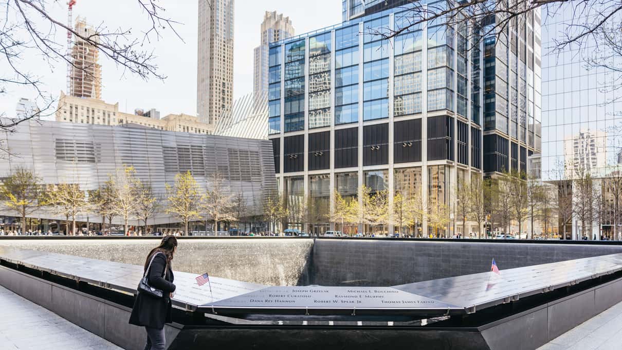 Visitor standing at the edge of a 9/11 Memorial pool with glass towers and the Oculus behind