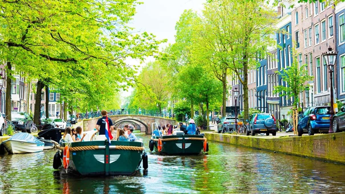 Barcos eléctricos verdes con pasajeros navegando por un canal bordeado de árboles y casas estrechas