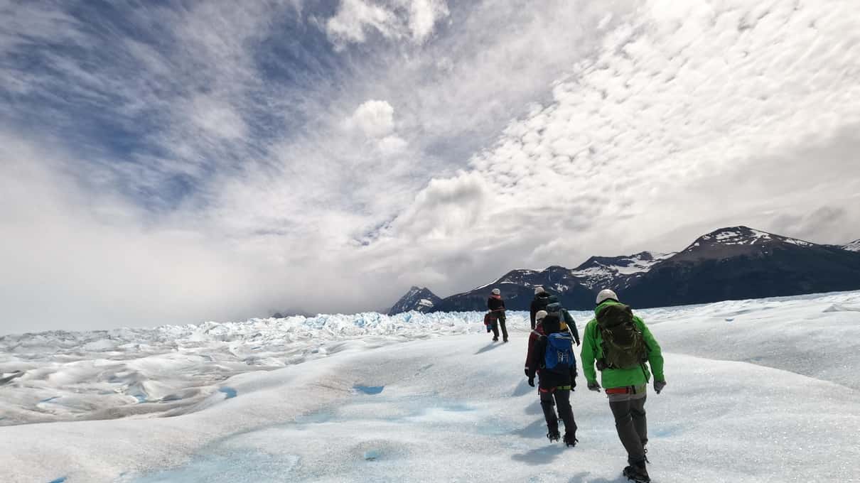 Hikers crossing a wide plain of Perito Moreno ice under a heavy gray sky with dark mountains