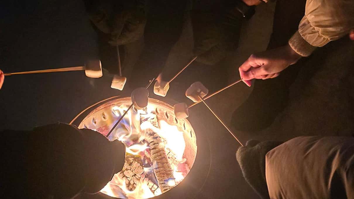 Guests in winter coats roasting marshmallows on sticks over a campfire bowl at night