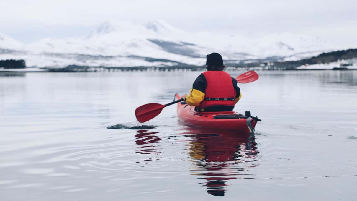 A kayaker in a red sea kayak paddling toward snowy mountains on a grey winter day
