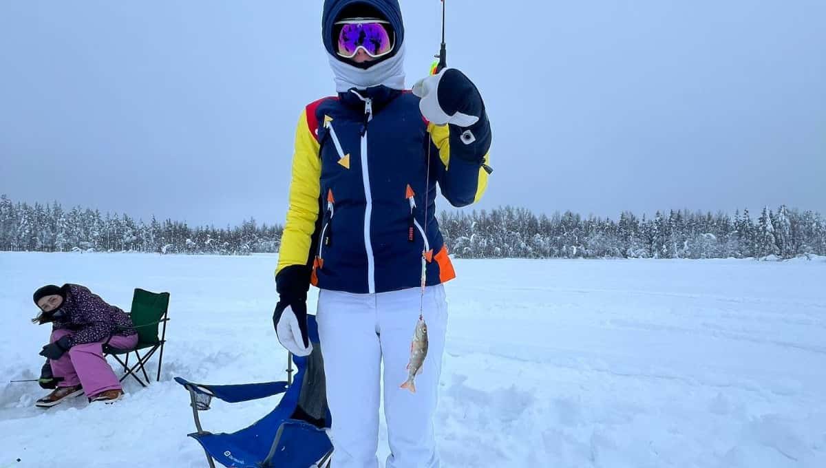 An angler in a bright winter suit holding up a small fish on a snowy frozen lake