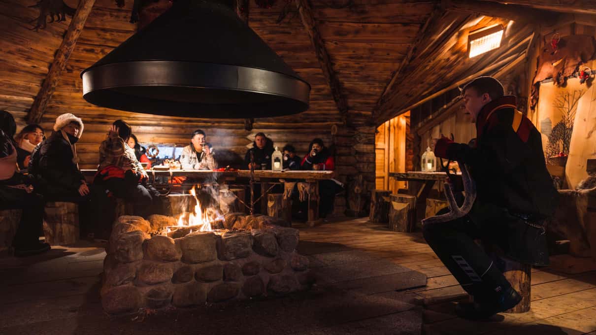 Guests in winter clothing sitting around an open fire inside a log kota hut