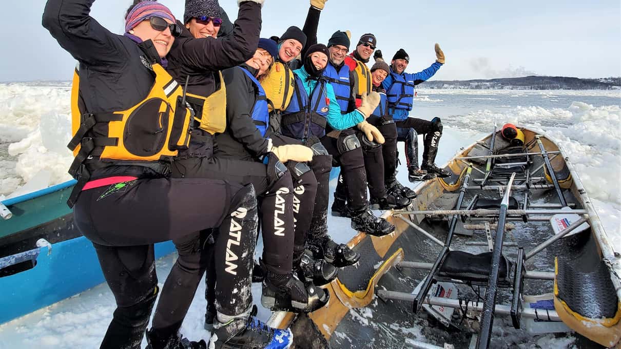 A crew in life jackets waving from an ice canoe among floating ice on the frozen St Lawrence