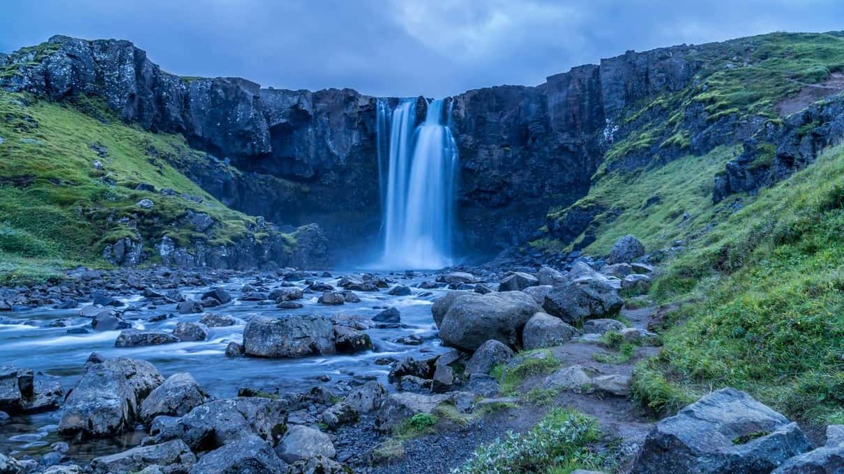Gufufoss waterfall pouring over a dark cliff into a rocky river under a blue sky near Seyðisfjörður