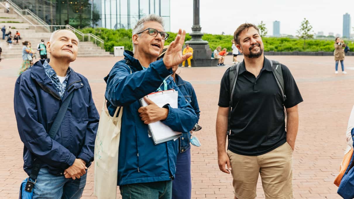 Guide holding a folder and pointing upward as two older men listen on the Liberty Island plaza, with the museum's glass building behind