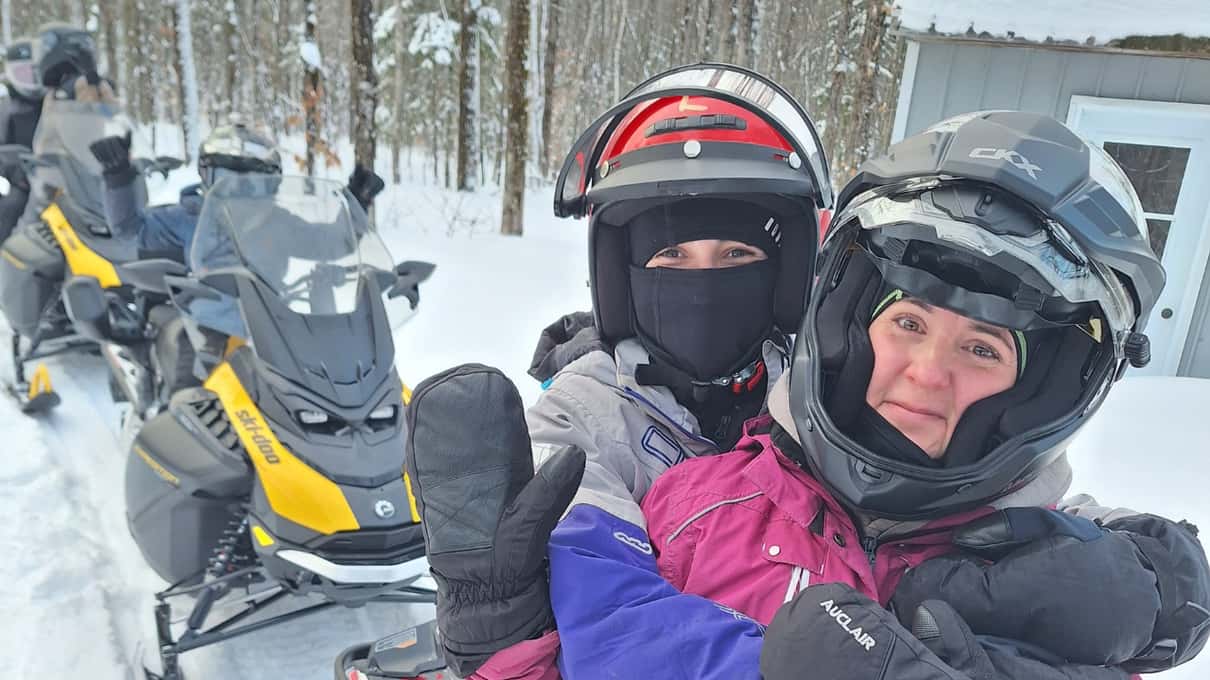 Two snowmobile riders in helmets and balaclavas taking a selfie beside their parked sleds