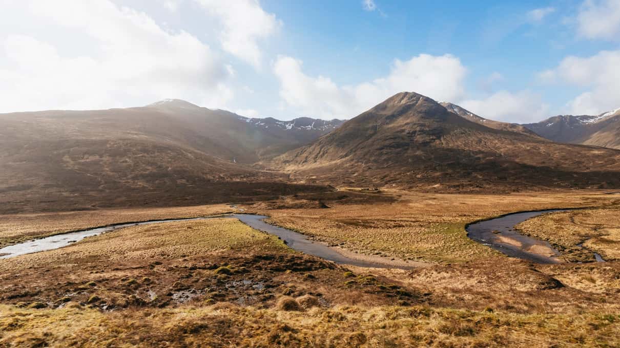 Bare brown moorland and a braided river below the snow-streaked slopes of the Red Cuillin on the Isle of Skye