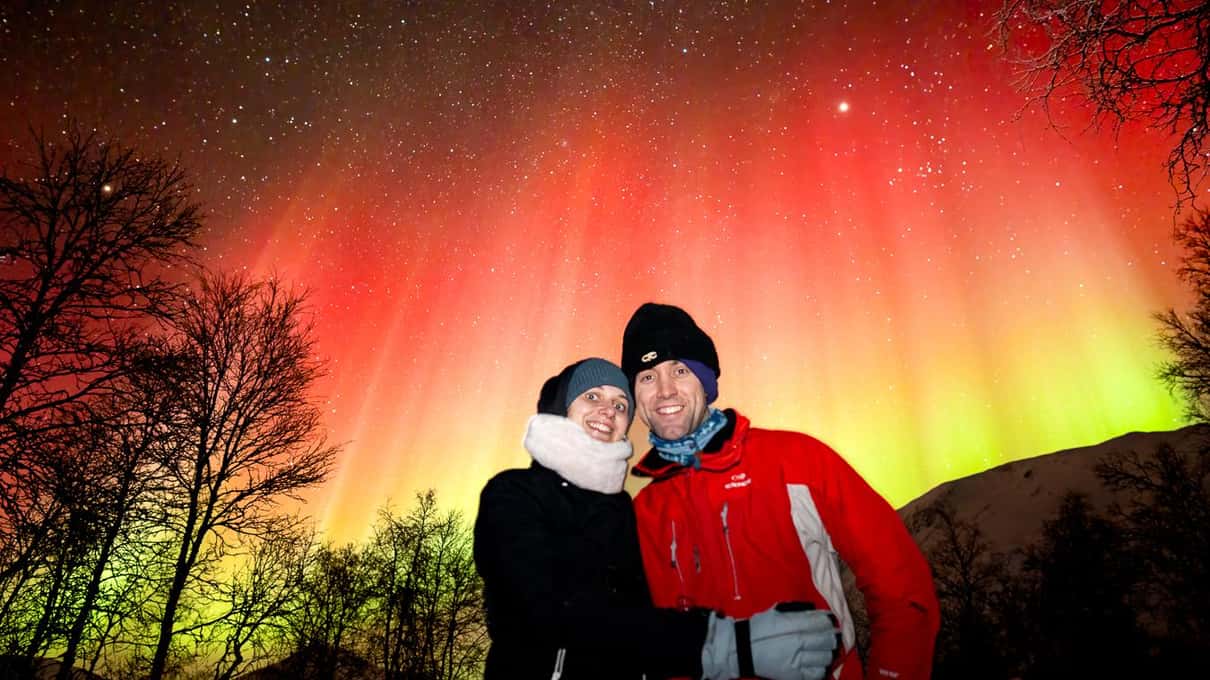 Smiling couple in winter jackets under a red and green aurora, framed by bare birch trees