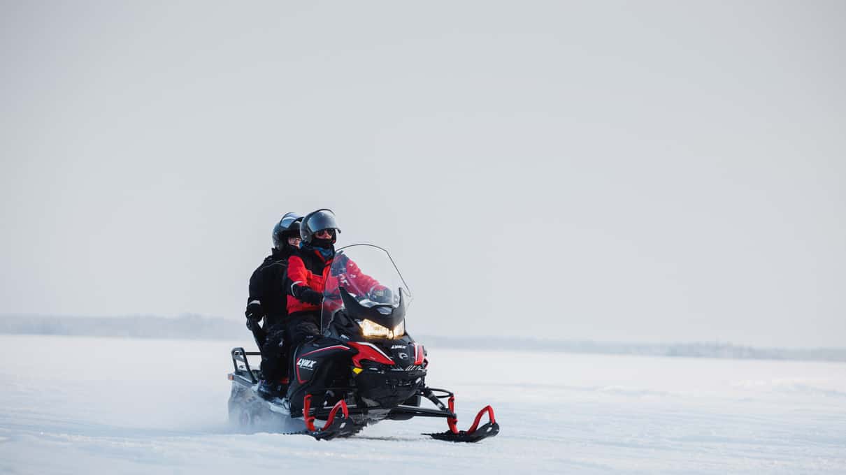 Two riders sharing one snowmobile on a wide, white frozen lake under a grey sky