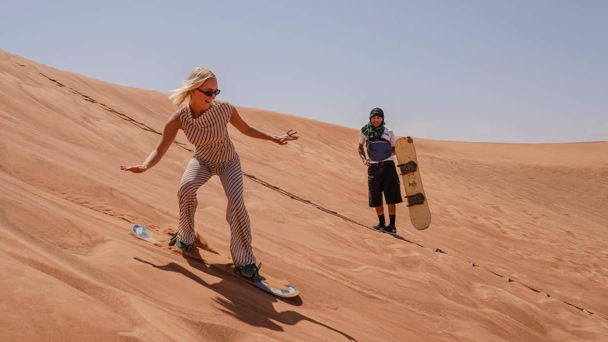 Una ragazza scende in sandboard lungo una duna rossa, dietro di lei un ragazzo con la tavola in mano