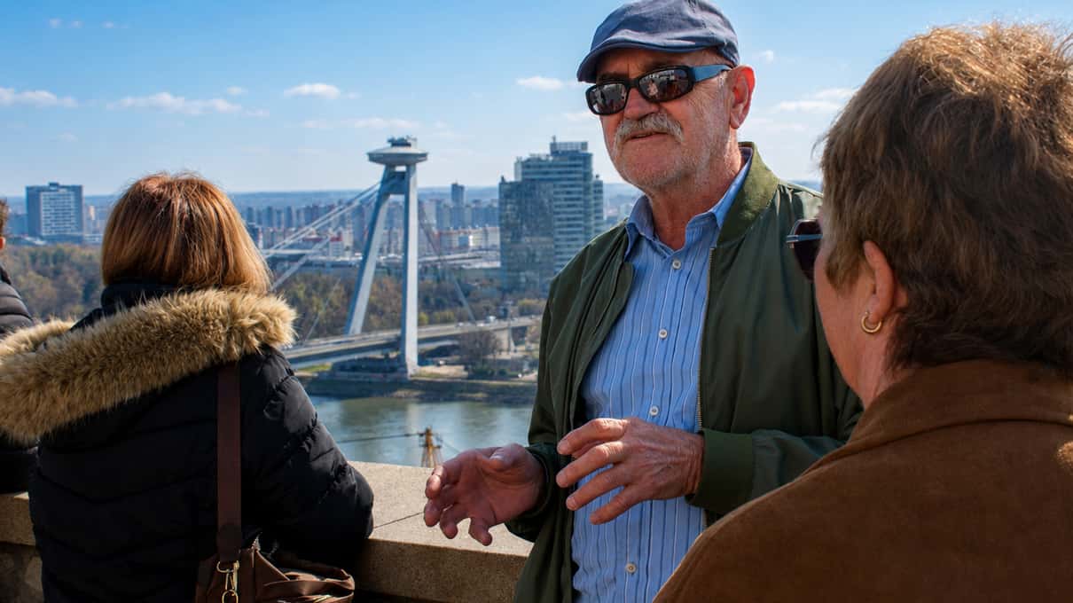 Travelers standing at a viewpoint high above the Danube in Bratislava, with the bridge and its round tower restaurant behind them