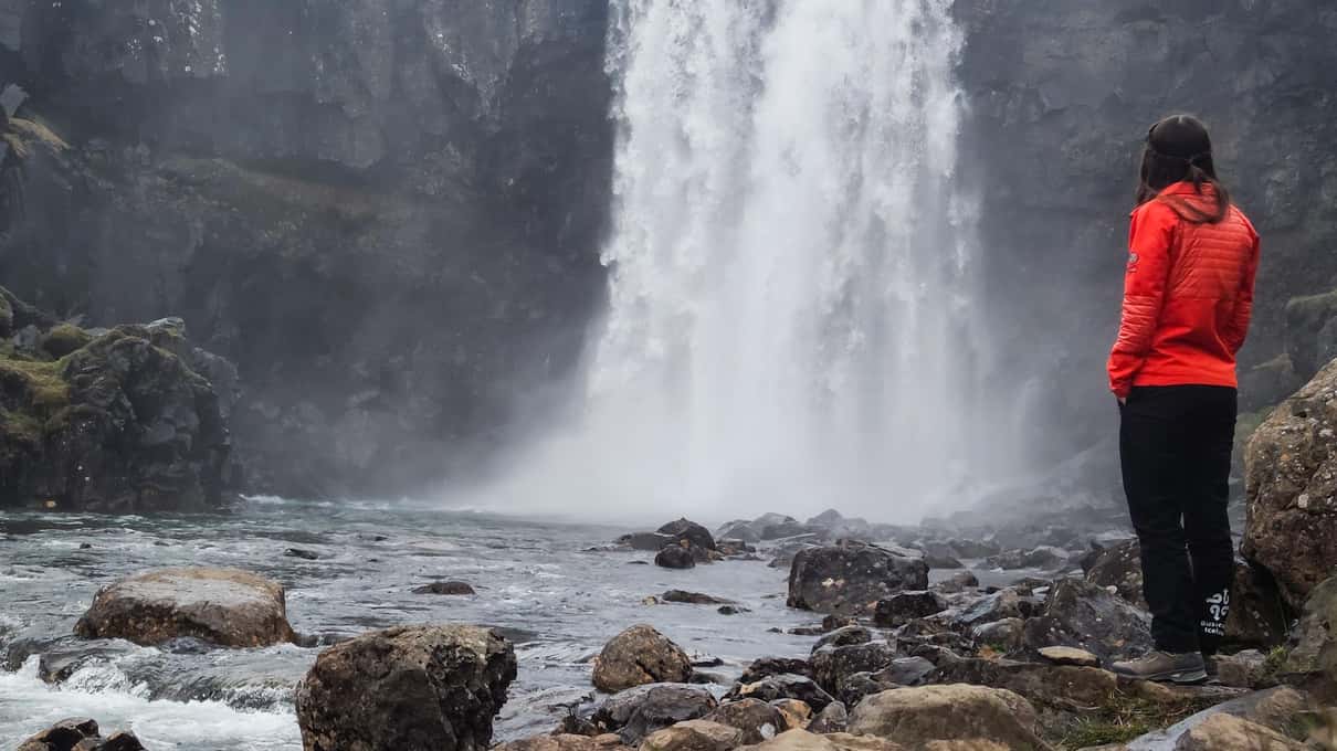 A visitor in a red jacket standing on the rocks below a powerful waterfall