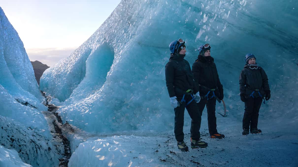 Three hikers in helmets and waterproof jackets walking between walls of blue glacier ice