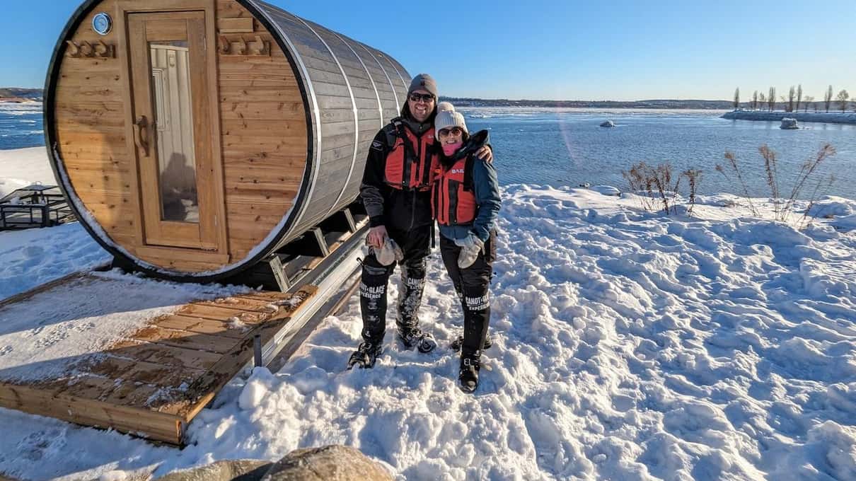 Two people in life vests standing beside a barrel sauna on the snowy St Lawrence shore