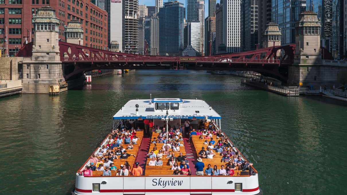 A sightseeing boat packed with passengers cruising down the middle of the Chicago River between rows of towers