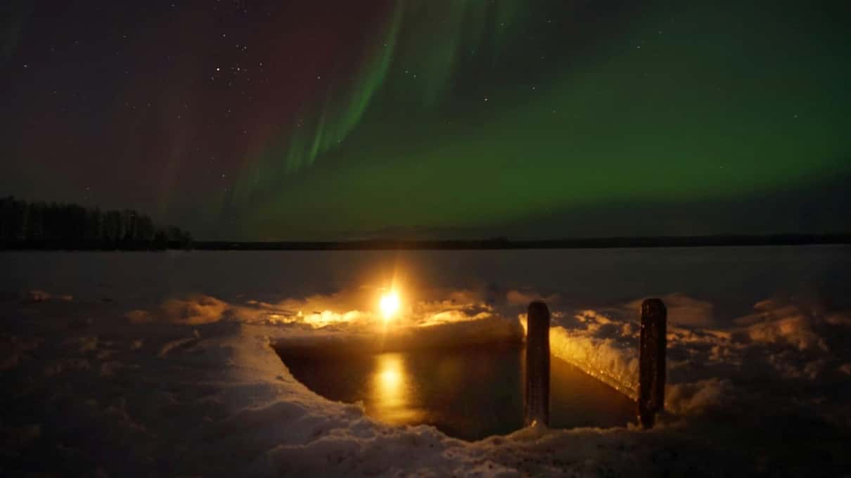 Green northern lights over the dark Kemijoki river with a small fire glowing on the bank by the sauna