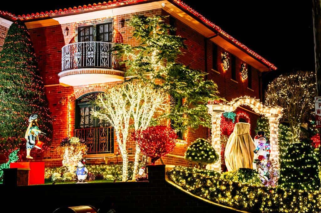 Bare street trees wrapped tightly in white lights in front of a house glowing red at night in Dyker Heights