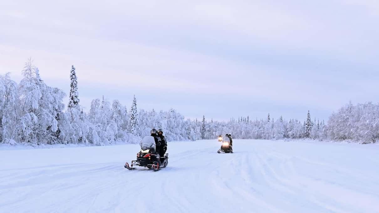 Two snowmobiles crossing a wide frozen lake with snowy forest on the far shore