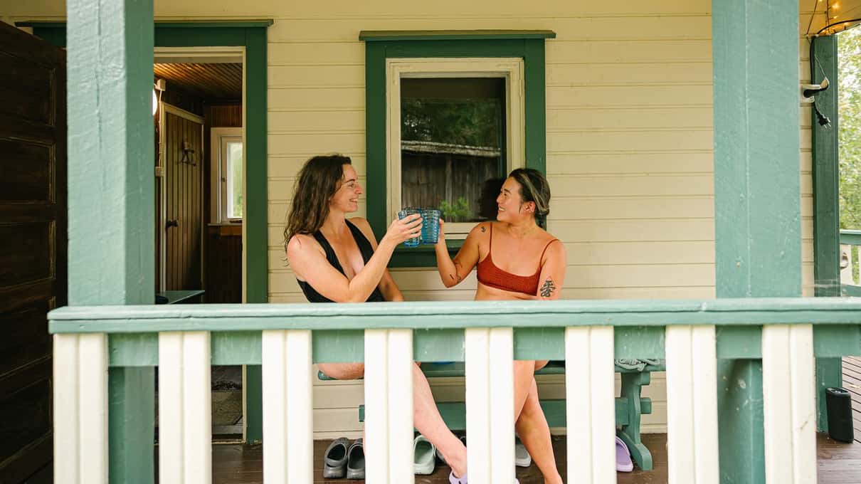 Two friends in swimsuits toasting with mugs on the wooden terrace of a Lapland sauna