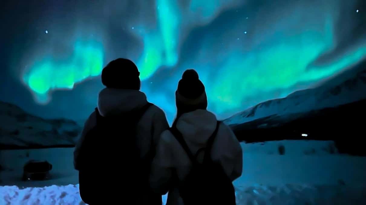Zwei Menschen von hinten als Silhouetten im Schnee, über ihnen ein leuchtend grünes Nordlicht