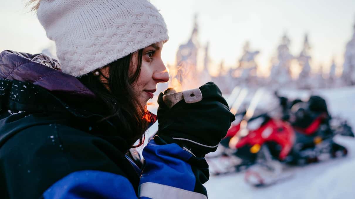 A rider in a white beanie and thermal suit holding a hot drink with snowmobiles parked behind
