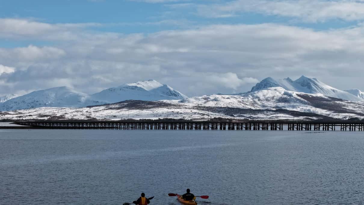 Two kayakers paddling toward a long low bridge with snowy mountains rising behind it
