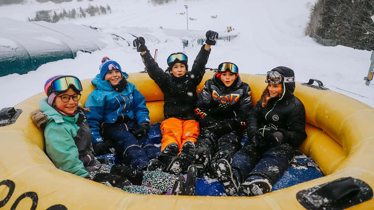 Five kids in goggles waving from a big yellow snow tube at Village Vacances Valcartier
