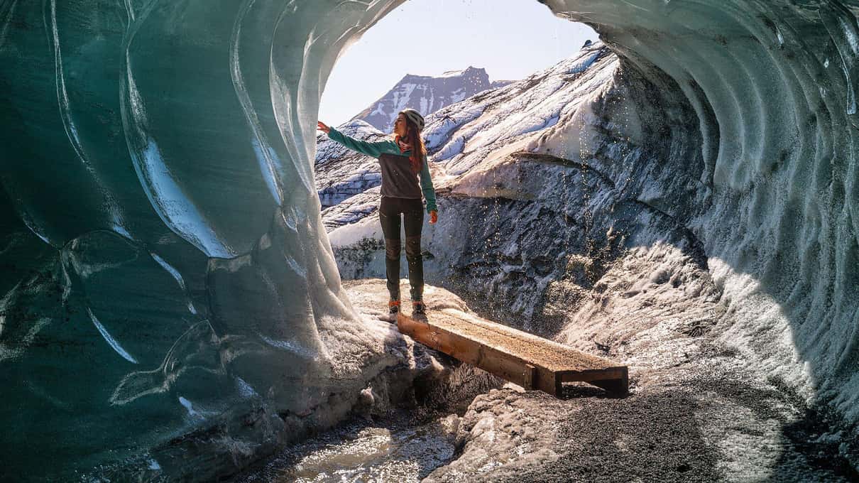 Un visiteur debout sur une planche de bois à l'entrée lumineuse de la grotte de glace de Katla, sous des arches de glace bleue et gris cendre