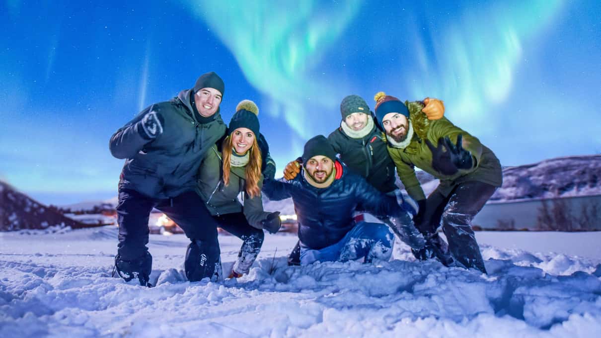 Fünf Freunde in Winterjacken knien lachend im Schnee, über ihnen ein grünes Nordlicht am blauen Nachthimmel