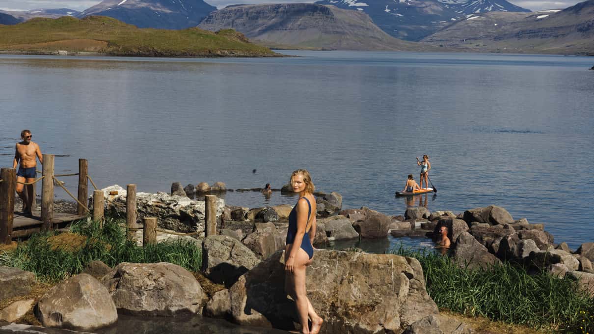 A bather sitting on dark rocks beside a hot pool with the fjord behind