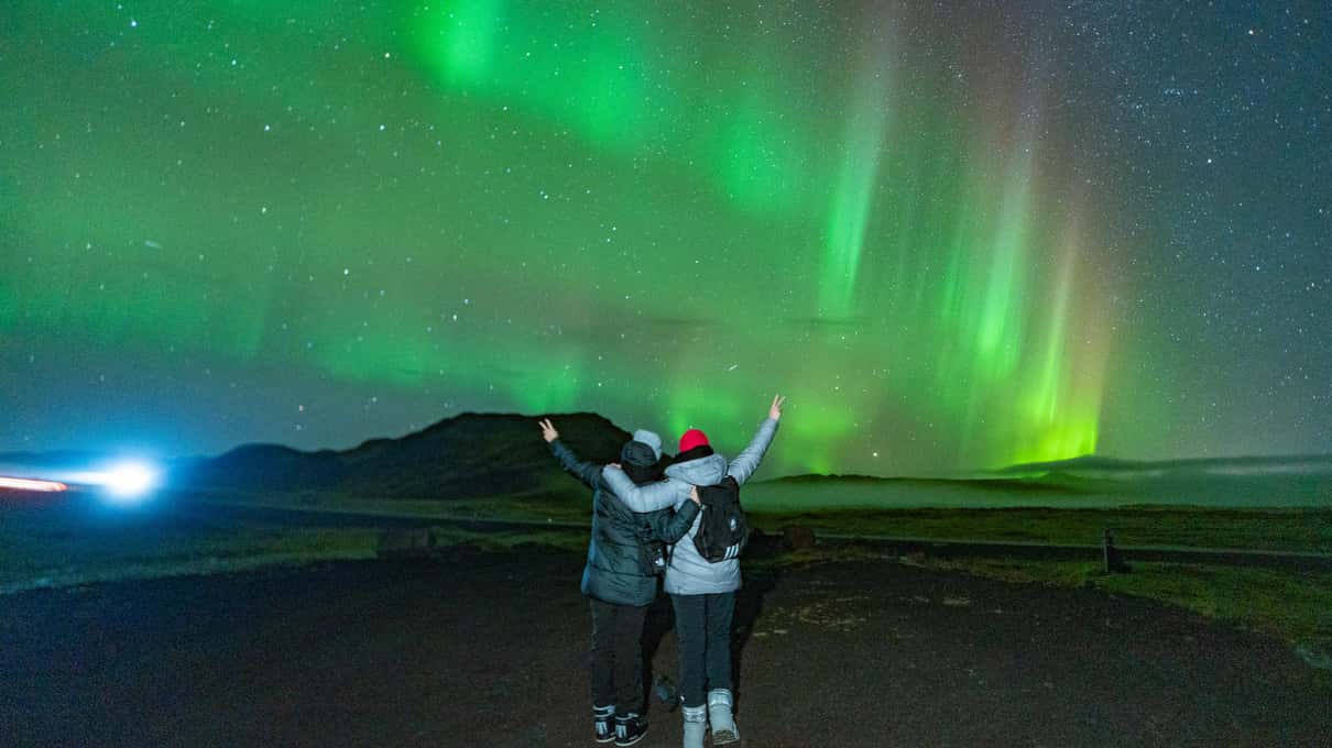 Two people standing under a green aurora while a head torch beam cuts across the dark ground