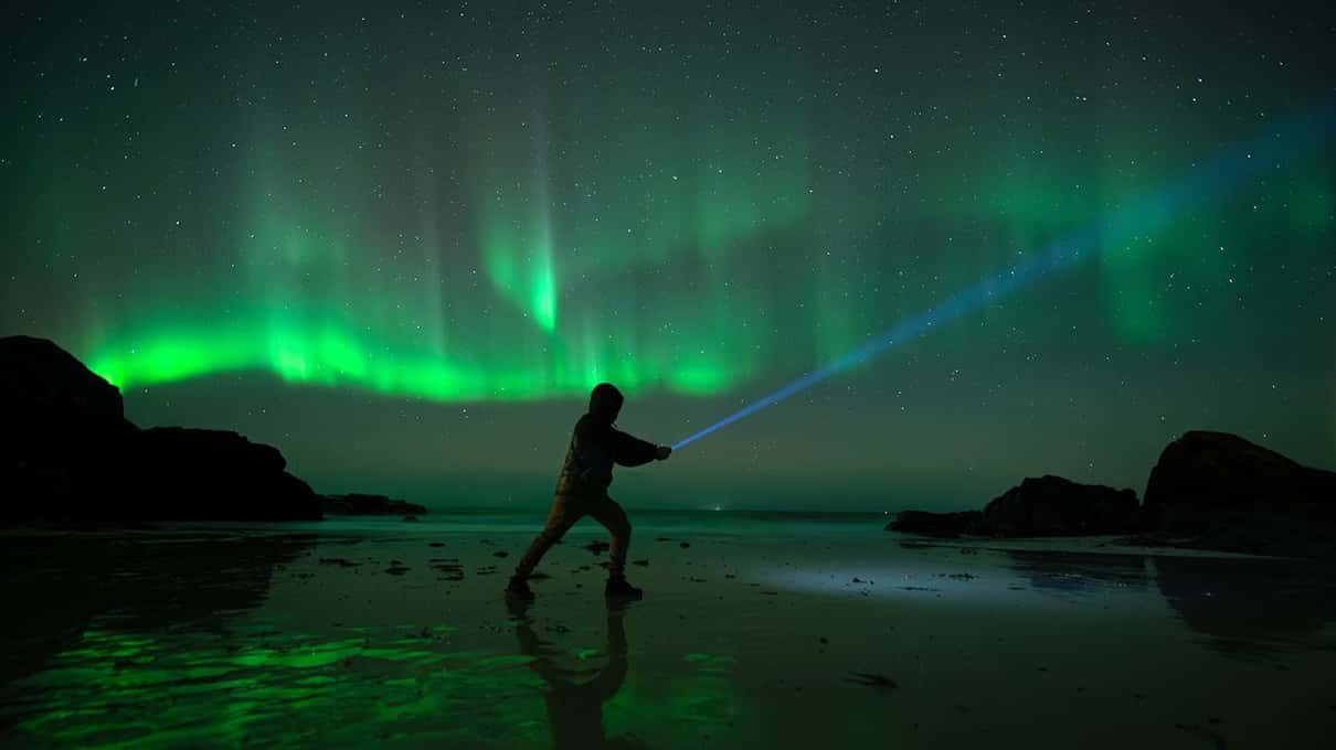A person standing on a dark beach between rocks, pointing a torch beam under a green aurora