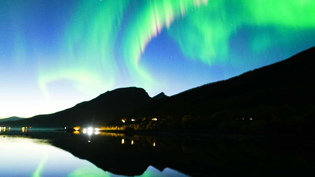 Grünes Nordlicht wölbt sich über einen Bergrücken und spiegelt sich in einem stillen See, am Ufer ein paar Lichter