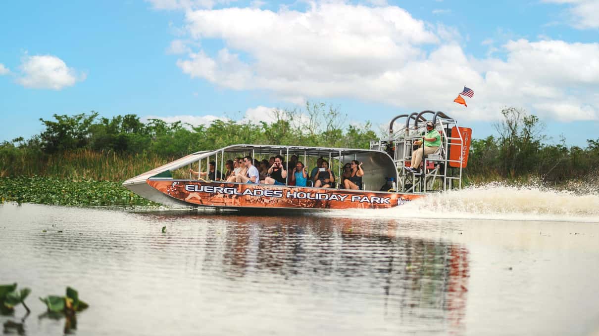 Red airboat full of riders crossing open marsh under a sky of white clouds