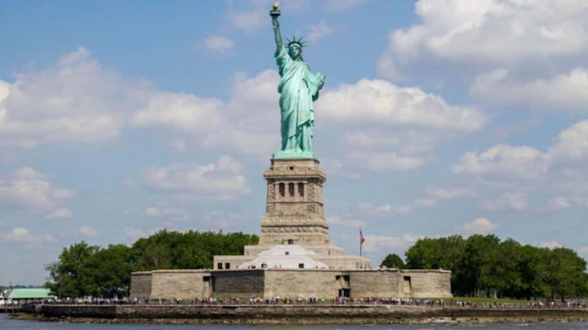 The Statue of Liberty and Liberty Island seen from the water under a blue sky with white clouds
