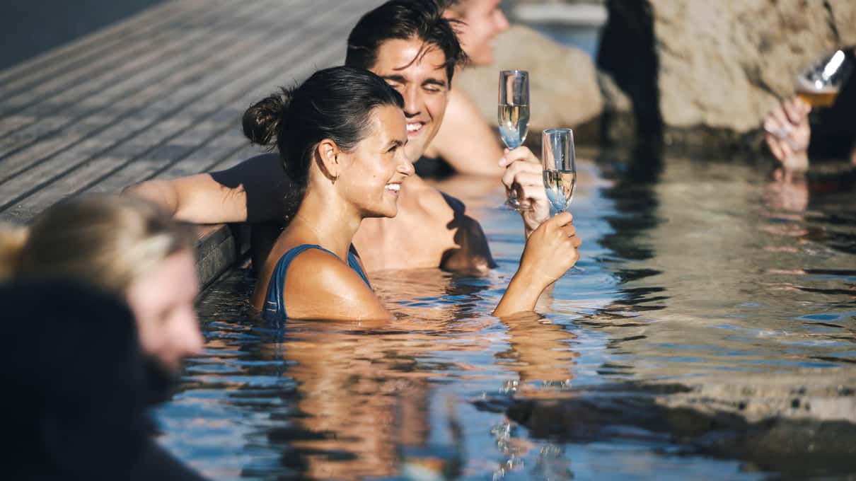 Two bathers raising glasses in a Hvammsvik pool with the fjord behind them
