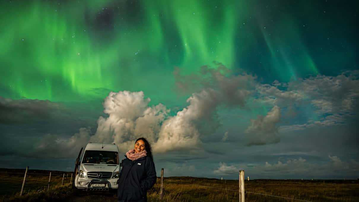 A white 4x4 minibus and a couple standing under a green aurora with low clouds near Reykjavík