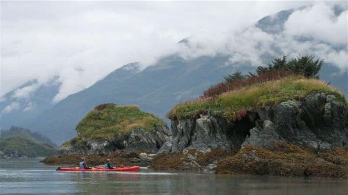 Two kayaks paddling past low grassy islets with cloud sitting halfway down the mountains behind