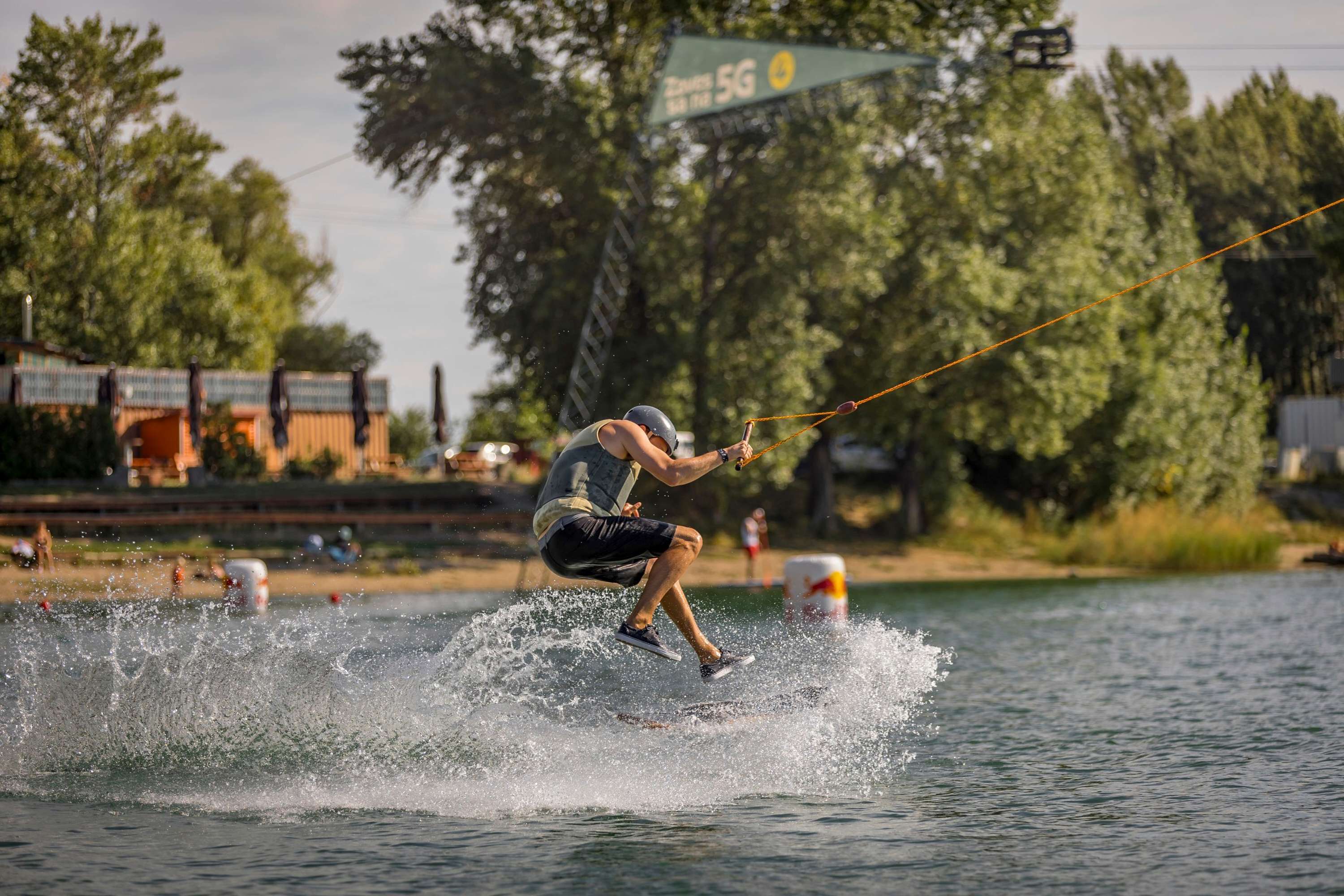 Wakeboarder performing a jump on a cable lake, sending water spraying into the air.