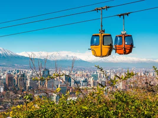 Two colorful gondolas on a cable car line with a view of Santiago, Chile, and the snow-capped Andes mountains in the background.