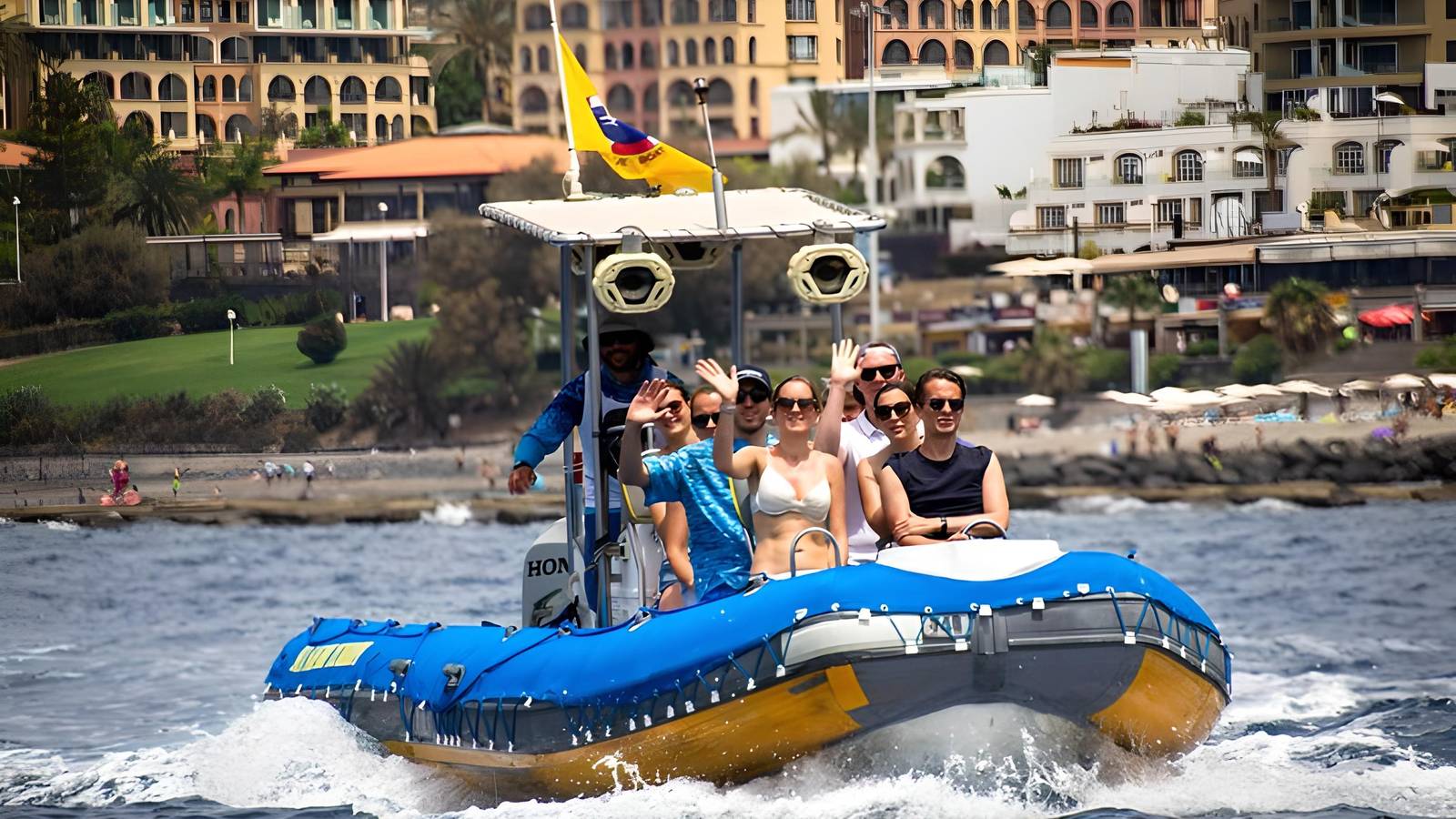 Blue RIB with passengers waving, Costa Adeje resort behind, Barco Azul flag on mast