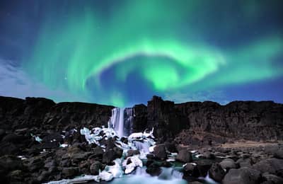 Green aurora swirl over a small waterfall between dark lava cliffs